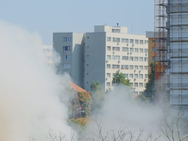 View of flames getting out of burning house in city landscape. Fumes and smog going out of building on fire while firefighters trying to extinguish dangerous smoke with water.